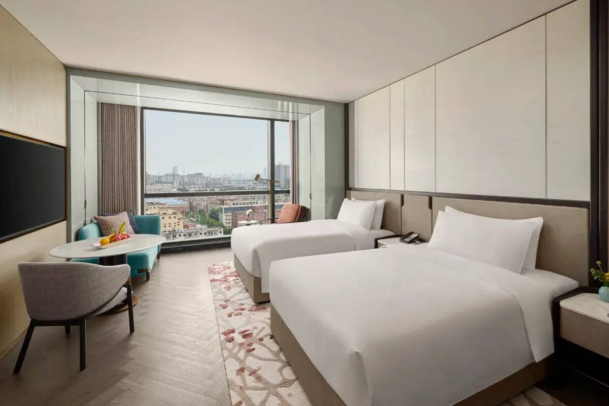 Een hotelkamer met twee bedden, een klein tafeltje, een stoel en een groot raam met uitzicht over de stad.