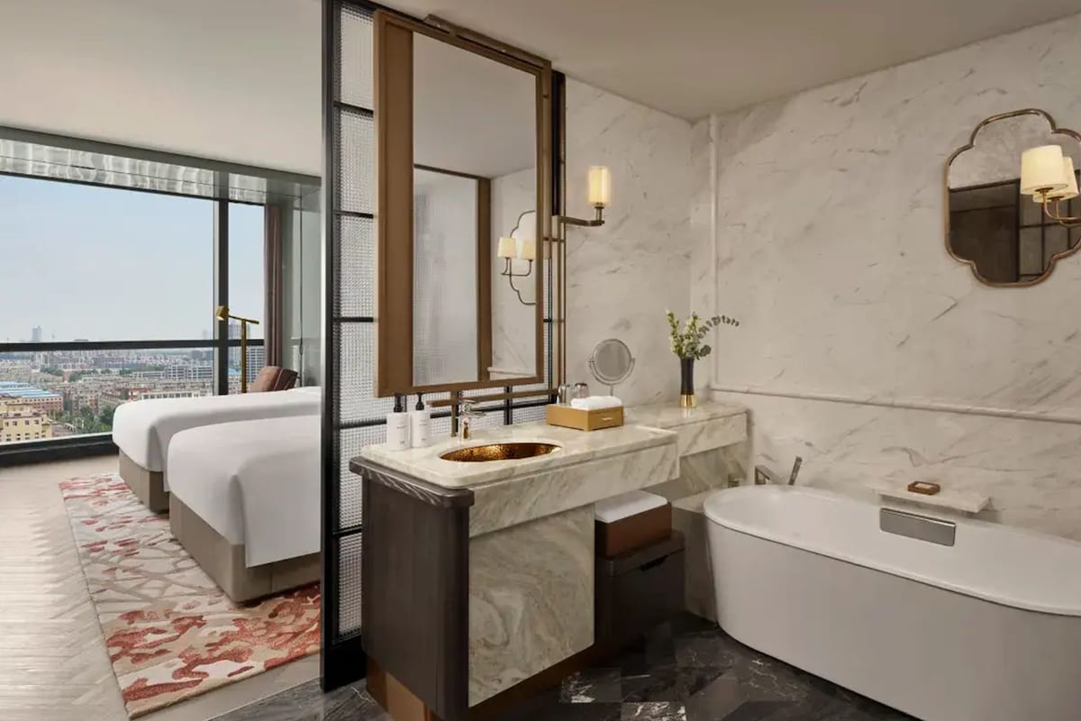 A modern hotel bathroom with a large mirror, a bathtub, and a sink. There is a large window providing a city view.