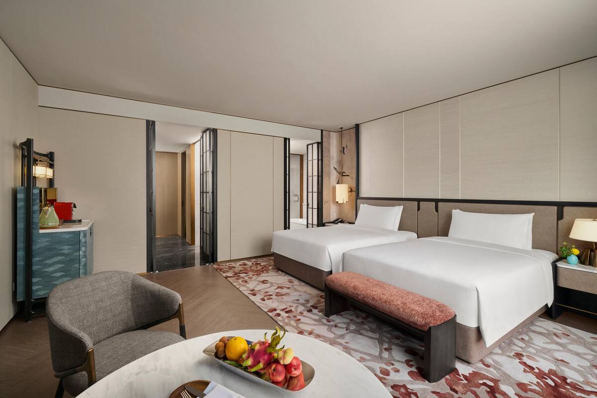 Een hotelkamer met twee bedden, een bureau, een stoel, een schaal fruit en een nachtkastje met een lamp.