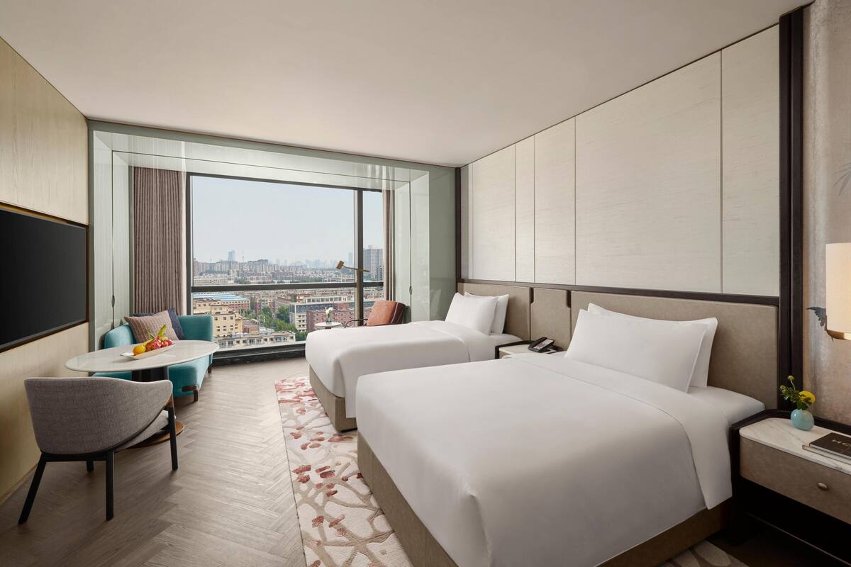 Een hotelkamer met twee bedden, een kleine tafel met stoelen, een televisie en uitzicht op de stad.