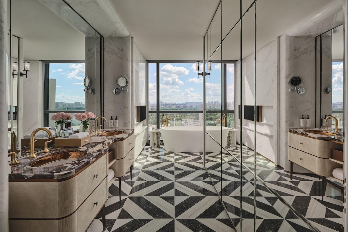 A modern bathroom with a large bathtub, two sinks, and a large mirror. The floor has a black and white geometric pattern.
