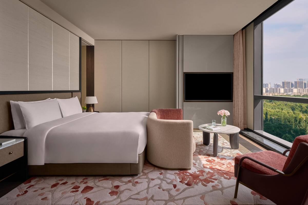 Een moderne hotelkamer met een groot bed, een zithoek met een stoel en een tafeltje, een aan de muur bevestigde televisie, en een stadszicht door grote ramen.