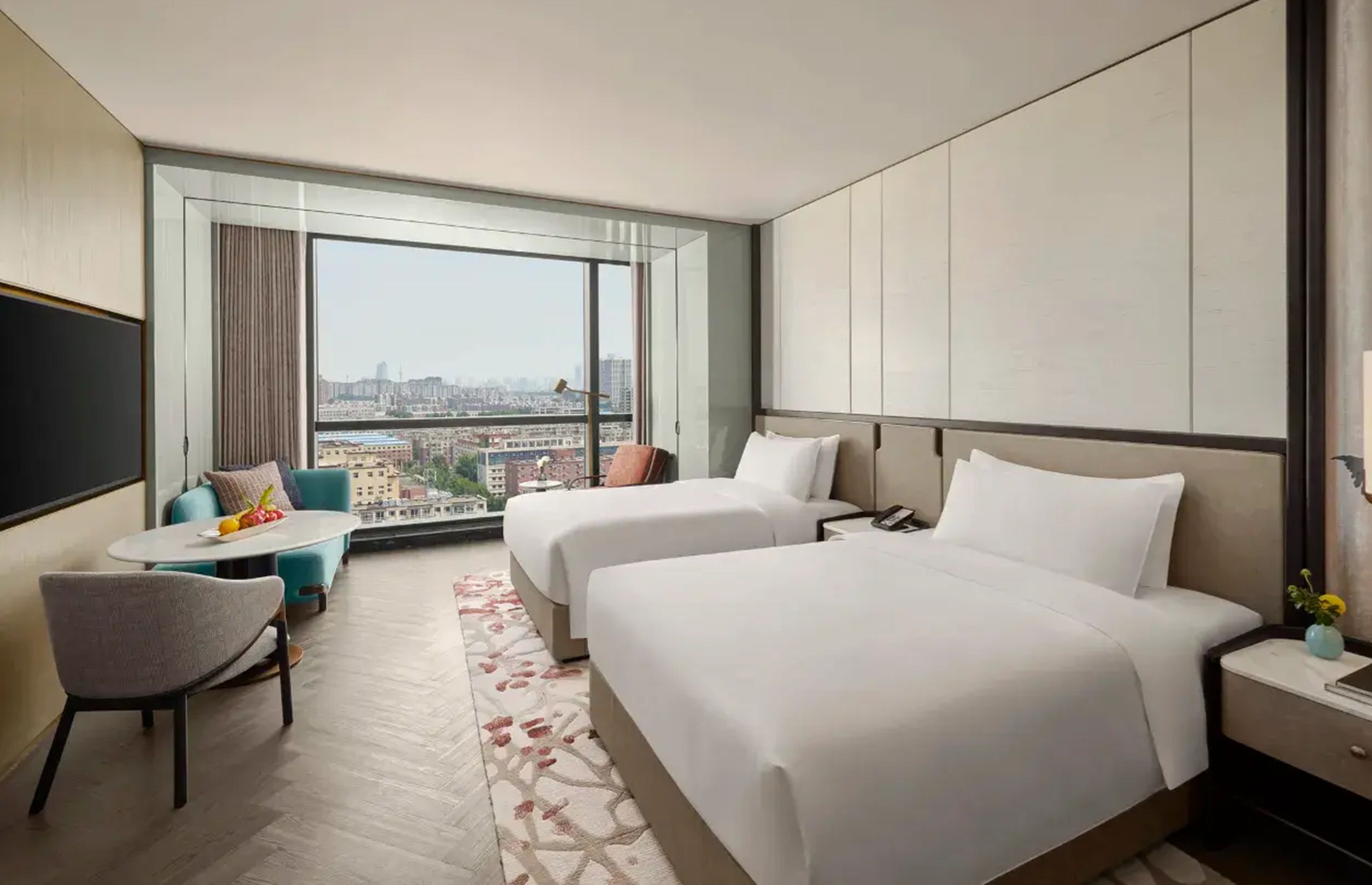 Ein Hotelzimmer mit zwei Betten, einem kleinen Tisch, einem Stuhl und einem großen Fenster mit Blick auf die Stadt.