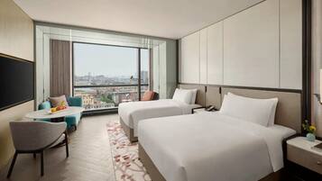 Ein Hotelzimmer mit zwei Betten, einem kleinen Tisch, einem Stuhl und einem großen Fenster mit Blick auf die Stadt.