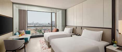 Habitación de hotel con dos camas, una mesita con sillas, un televisor y vistas a la ciudad.