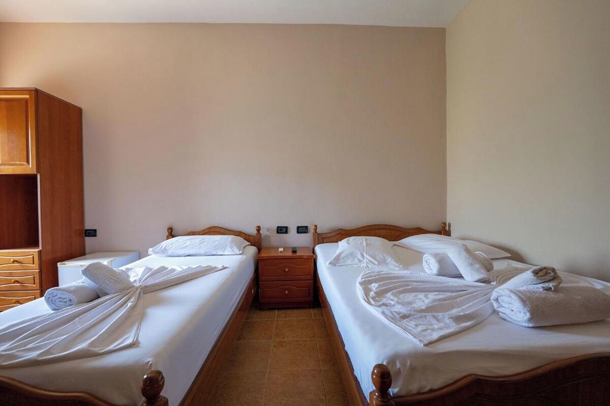 A hotel room with two single beds, wooden headboards, a wooden wardrobe, and a small bedside table with a lamp.