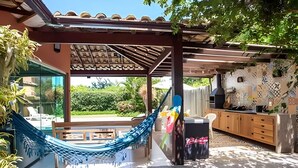 Um alpendre coberto com uma rede, espaço para churrasco e vista para uma piscina e vegetação.