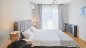 A modern bedroom with a large bed, a bedside table with a lamp, a radiator, and a window with sheer curtains. - Hotel White House (Tbilisi)