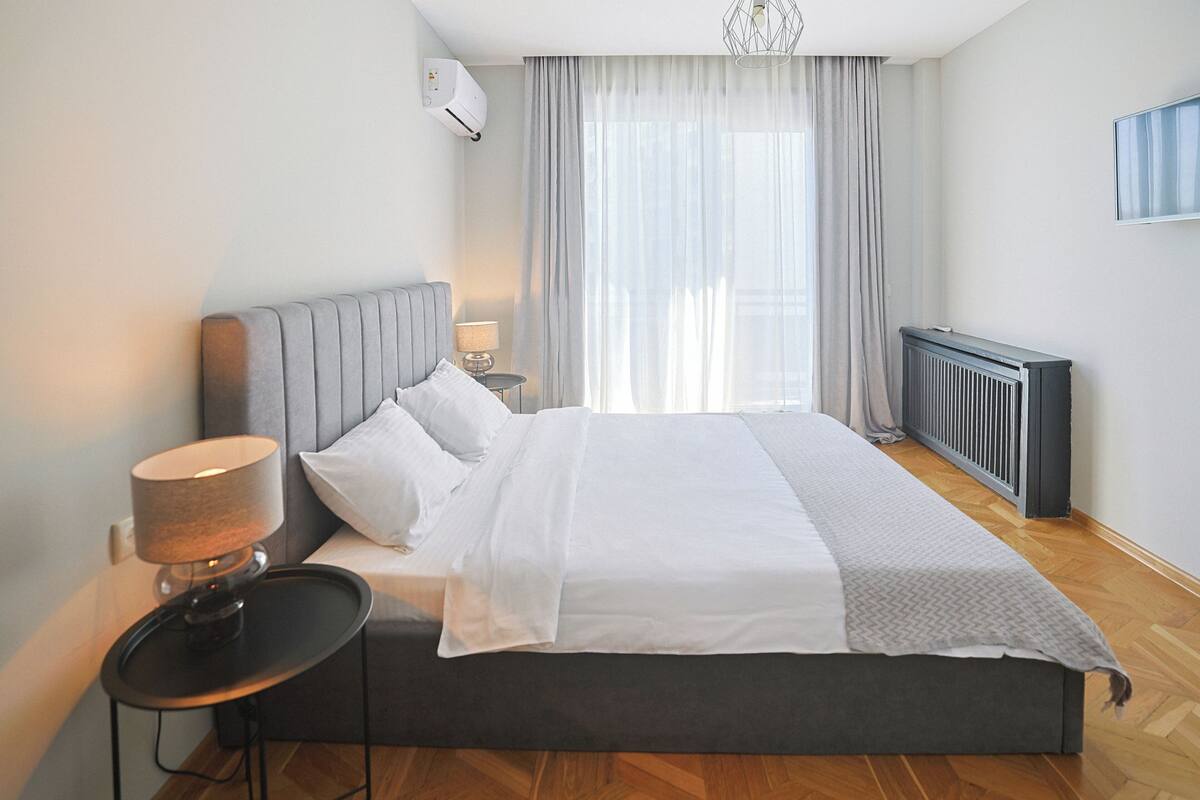 A modern bedroom with a large bed, a bedside table with a lamp, a radiator, and a window with sheer curtains.