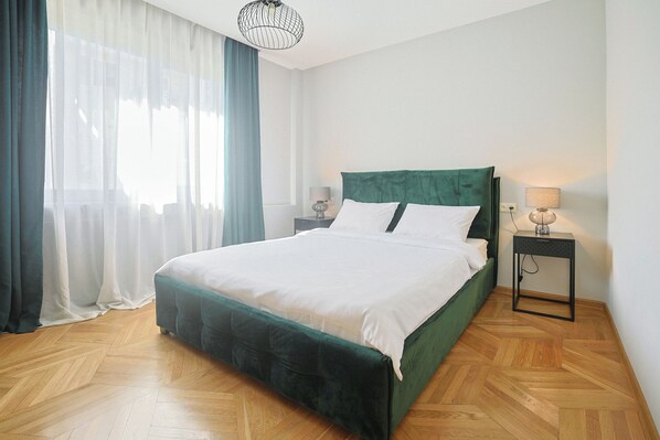 Une chambre à coucher bien aménagée, avec un lit recouvert d’un tissu vert, un parquet en bois disposé en chevrons, une table de chevet avec une lampe et une fenêtre ornée de rideaux légers.
