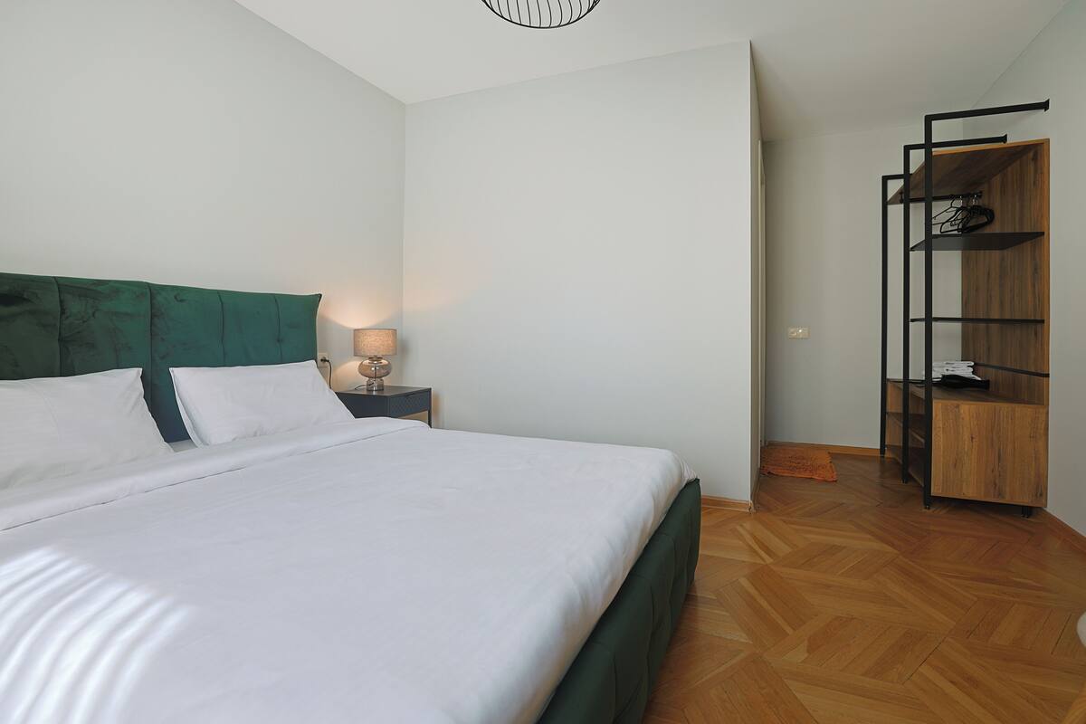 A neatly made bed with white linens, a green upholstered headboard, and a bedside table with a lamp.