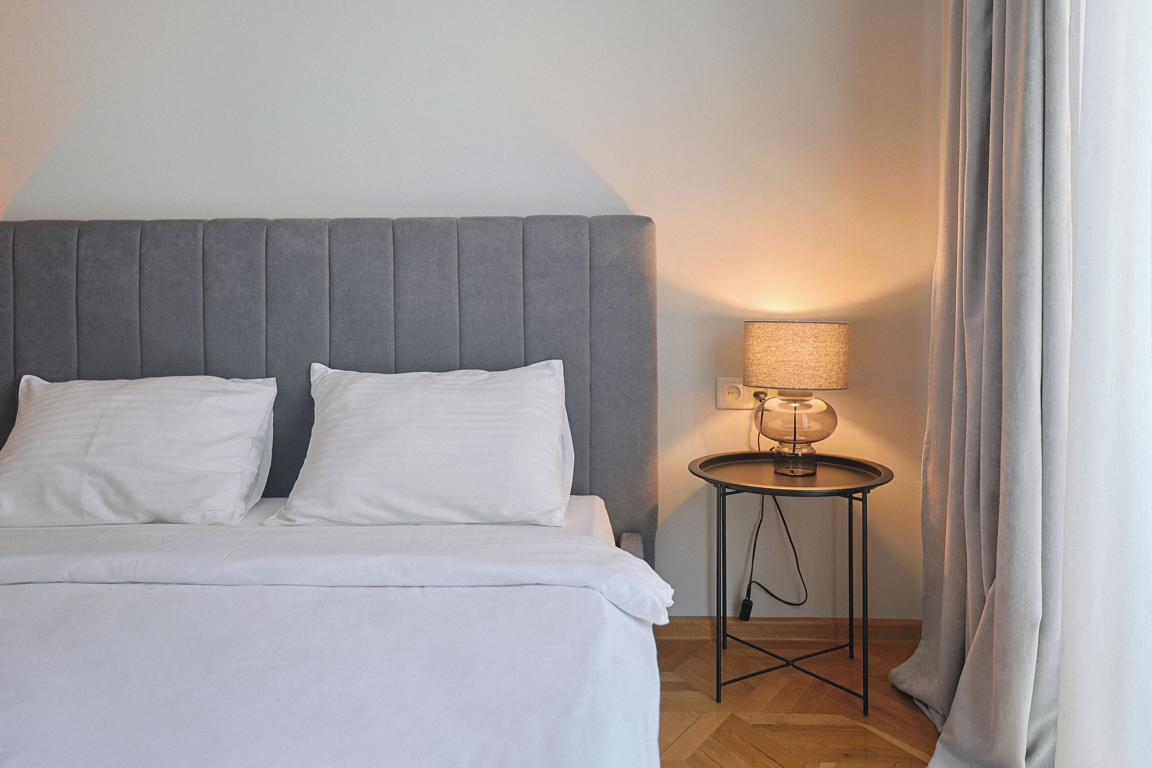 A neatly made bed with white linens, a grey upholstered headboard, and a small round table with a lamp beside it.