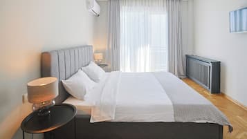A modern bedroom with a large bed, a bedside table with a lamp, a radiator, and a window with sheer curtains.