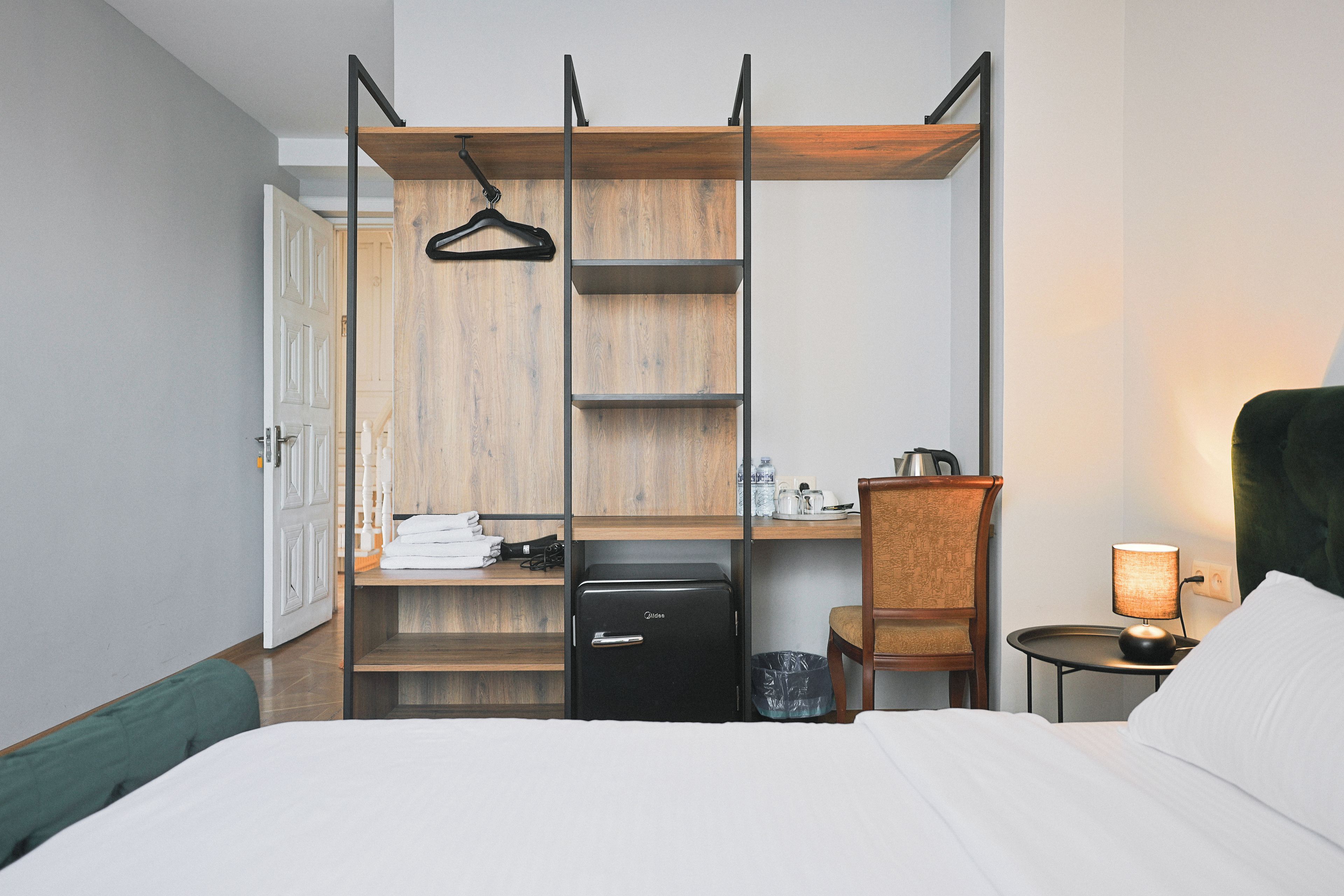 A hotel room with a bed, a desk with a chair, a wardrobe with shelves, a small black refrigerator, and a bedside table with a lamp.