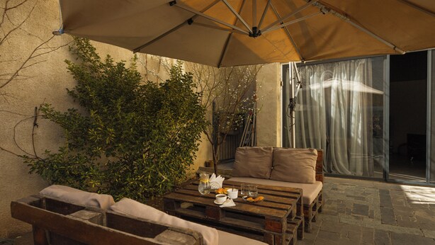 An outdoor seating area with wooden furniture and a table, a large umbrella providing shade, and a view of a building through a glass door. - Hotel White House (Tbilisi)