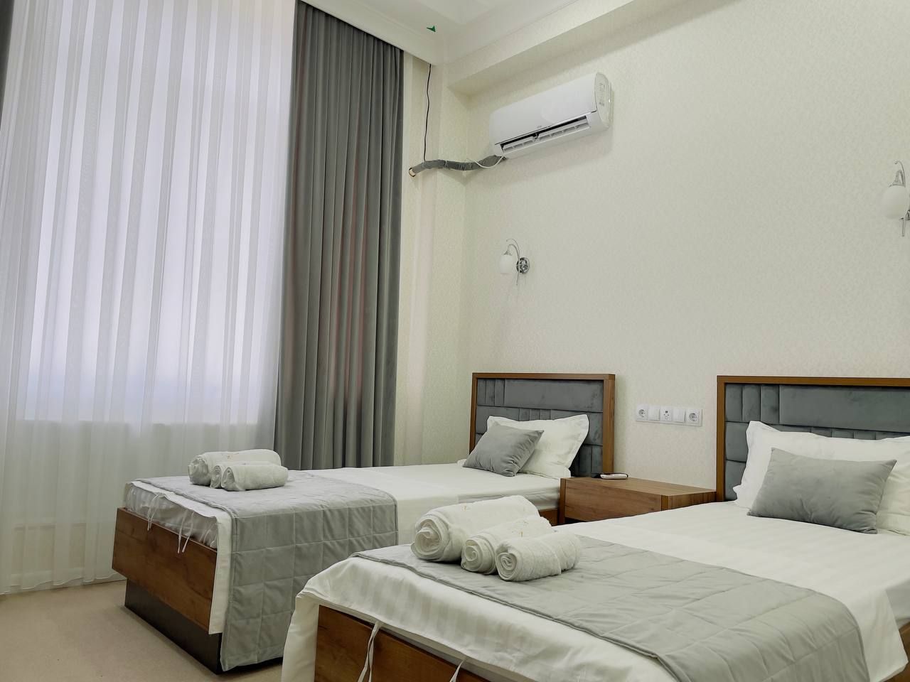 A hotel room with two single beds, a large window with sheer curtains, an air conditioning unit on the wall, and a small bedside table with a lamp.