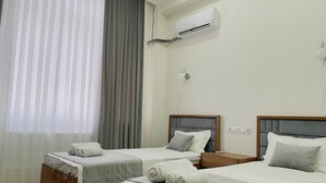 A hotel room with two single beds, a large window with sheer curtains, an air conditioning unit on the wall, and a small bedside table with a lamp. - Emin Hotel (Margilan)
