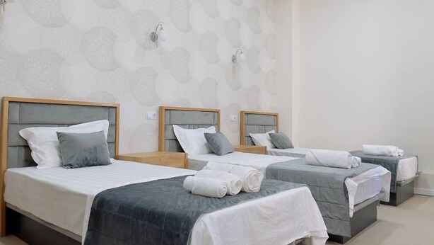 A hotel room with three beds, each with a headboard, white linens, and grey bedding. There are wall-mounted light fixtures and a patterned wallpaper. - Emin Hotel (Margilan)
