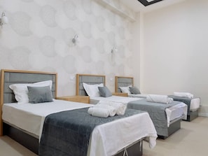 Una habitación de hotel con tres camas, cada una con cabecera, sábanas blancas y ropa de cama gris. Hay luminarias fijadas a la pared y un papel tapiz estampado.
