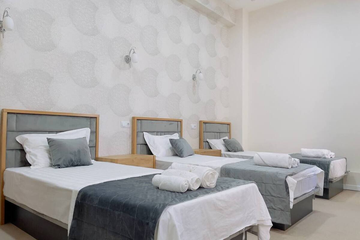 A hotel room with three beds, each with a headboard, white linens, and grey bedding. There are wall-mounted light fixtures and a patterned wallpaper.