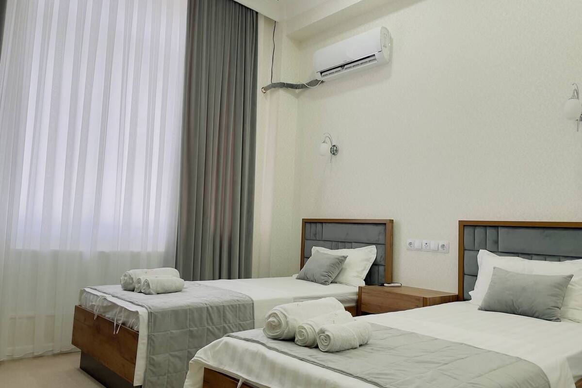 A hotel room with two single beds, a large window with sheer curtains, an air conditioning unit on the wall, and a small bedside table with a lamp.