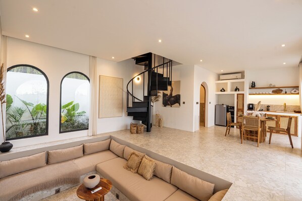 Un vaste espace de vie agrémenté d’un escalier noir aux lignes courbes, d’un grand canapé beige et d’un coin repas avec des chaises en bois.