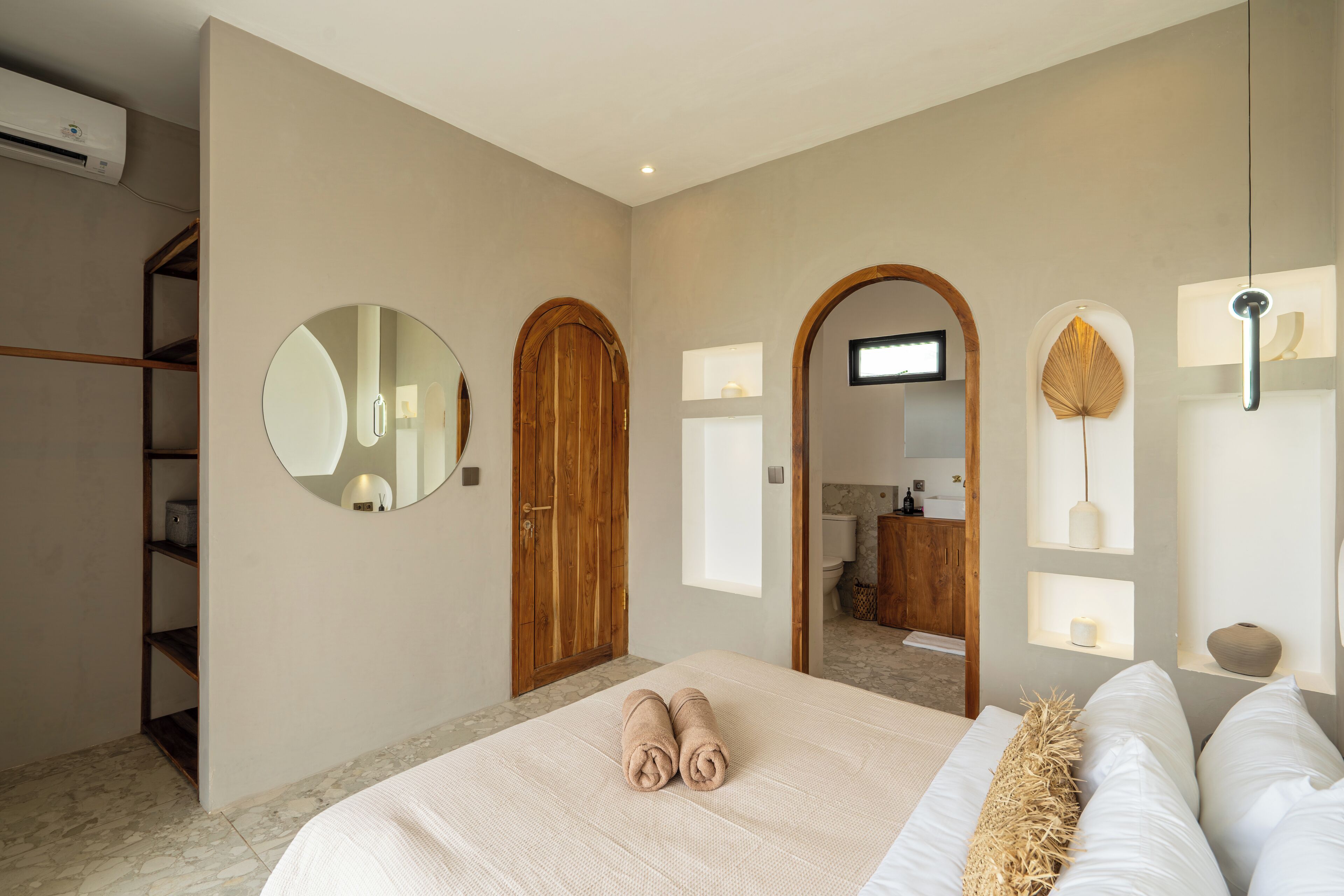 Une chambre à coucher avec un grand lit, une armoire en bois, un miroir rond et une salle de bain visible à travers une arche.