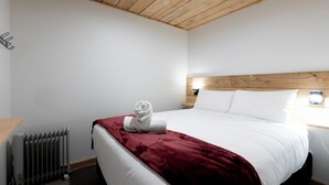 A hotel room with a wooden ceiling, a radiator, a neatly made bed with white bedding and a red blanket, and a wooden nightstand with a lamp. - Central Lofts Unit 3 (Queenstown)
