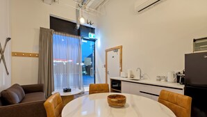 A modern hotel room with a round dining table, a sofa, a kitchen area with a refrigerator, and a view of the outside through a large window. - Central Lofts Unit 3 (Queenstown)