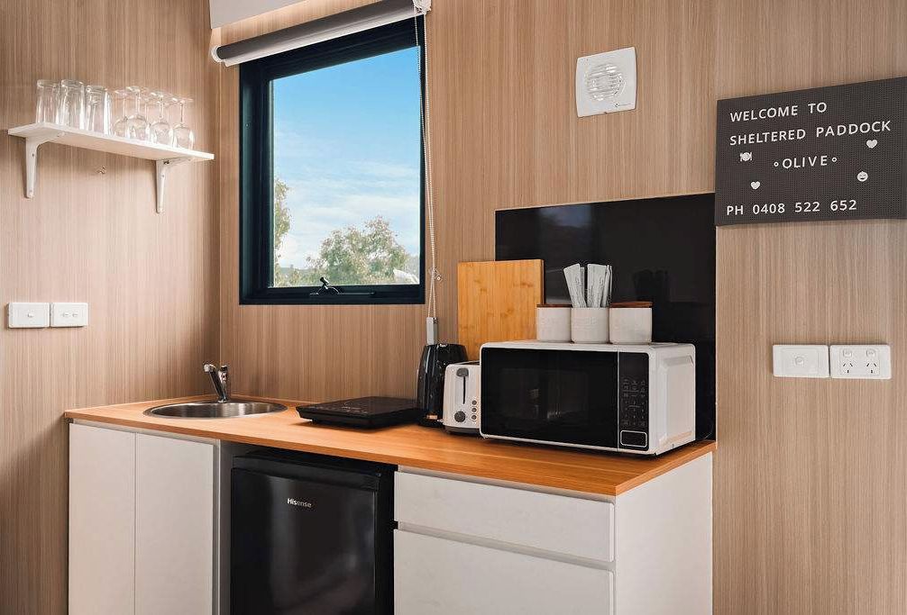 A compact kitchen with a wooden countertop, a sink, a microwave, and a toaster. A welcome sign and a window are also visible.