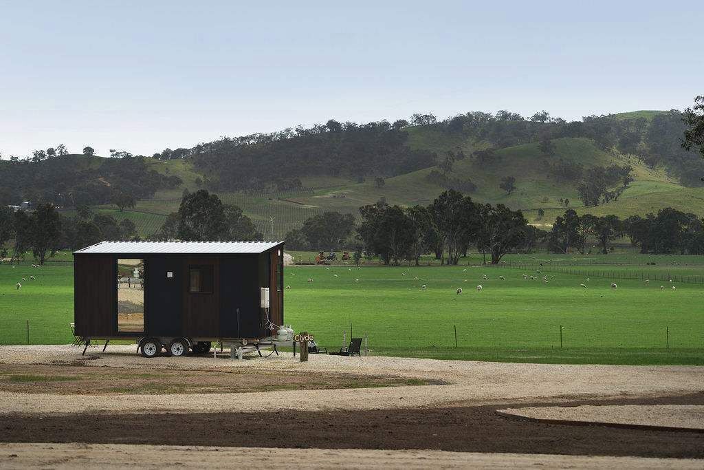 A small, dark-colored mobile home with a metal roof, situated on a gravel area with a green field and trees in the background.