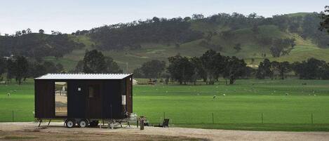 Una casa móvil pequeña y de color oscuro con techo de metal, ubicada en un área de grava con un campo verde y árboles al fondo.