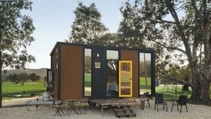 Une cabane moderne et compacte, dotée d’une façade en bois et de grandes fenêtres, située dans un espace verdoyant, agrémenté d’arbres et offrant une vue sur un ciel dégagé.