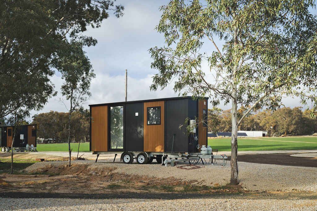 Una pequeña cabaña moderna con un amplio ventanal corredera, ubicada sobre una zona de grava con árboles y un campo de césped al fondo.
