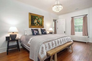 A bedroom with a bed, two bedside tables with lamps, a bench, a chandelier, a door, and a window with curtains. - Prime Location, Recently Redesigned, Riverfront! (Great Barrington)