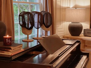 A grand piano, a stack of books, a lit candle, and three standing fans in a room with a window and a lamp. - Prime Location, Recently Redesigned, Riverfront! (Great Barrington)