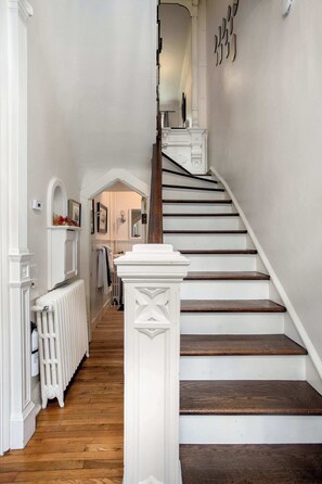 A staircase with white railings and dark wooden steps, leading to a landing with a radiator and a white wall with a mirror and framed pictures. - Prime Location, Recently Redesigned, Riverfront! (Great Barrington)