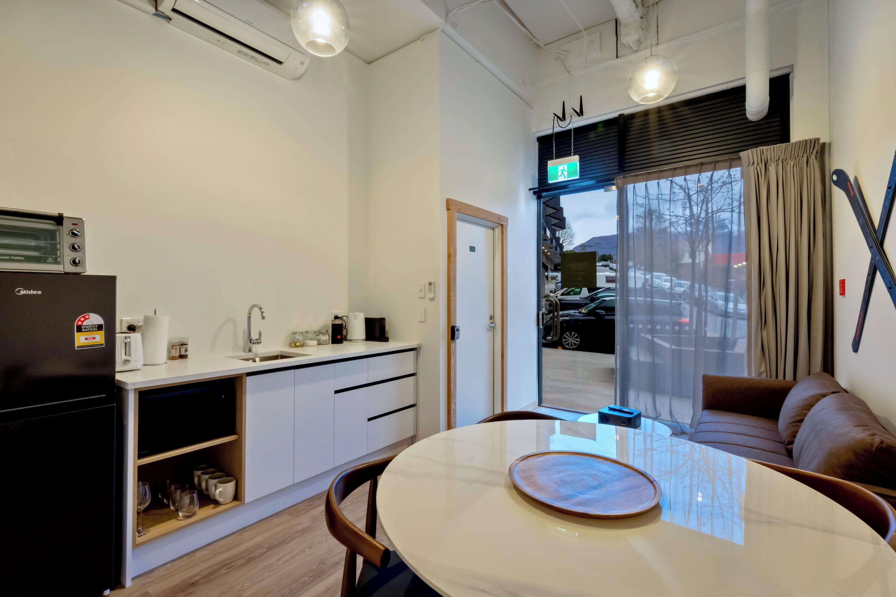 Une cuisine moderne équipée d’un réfrigérateur, d’un évier et d’un plan de travail. On y trouve également un coin repas avec une table et des chaises. Une fenêtre offre une vue sur la rue et les voitures qui passent.