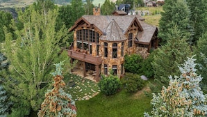 A large stone house with multiple windows and a balcony, surrounded by lush greenery and mountains in the background. - Luxe Mountain Village Retreat | Hot Tub + Shuttle (Mountain Village)