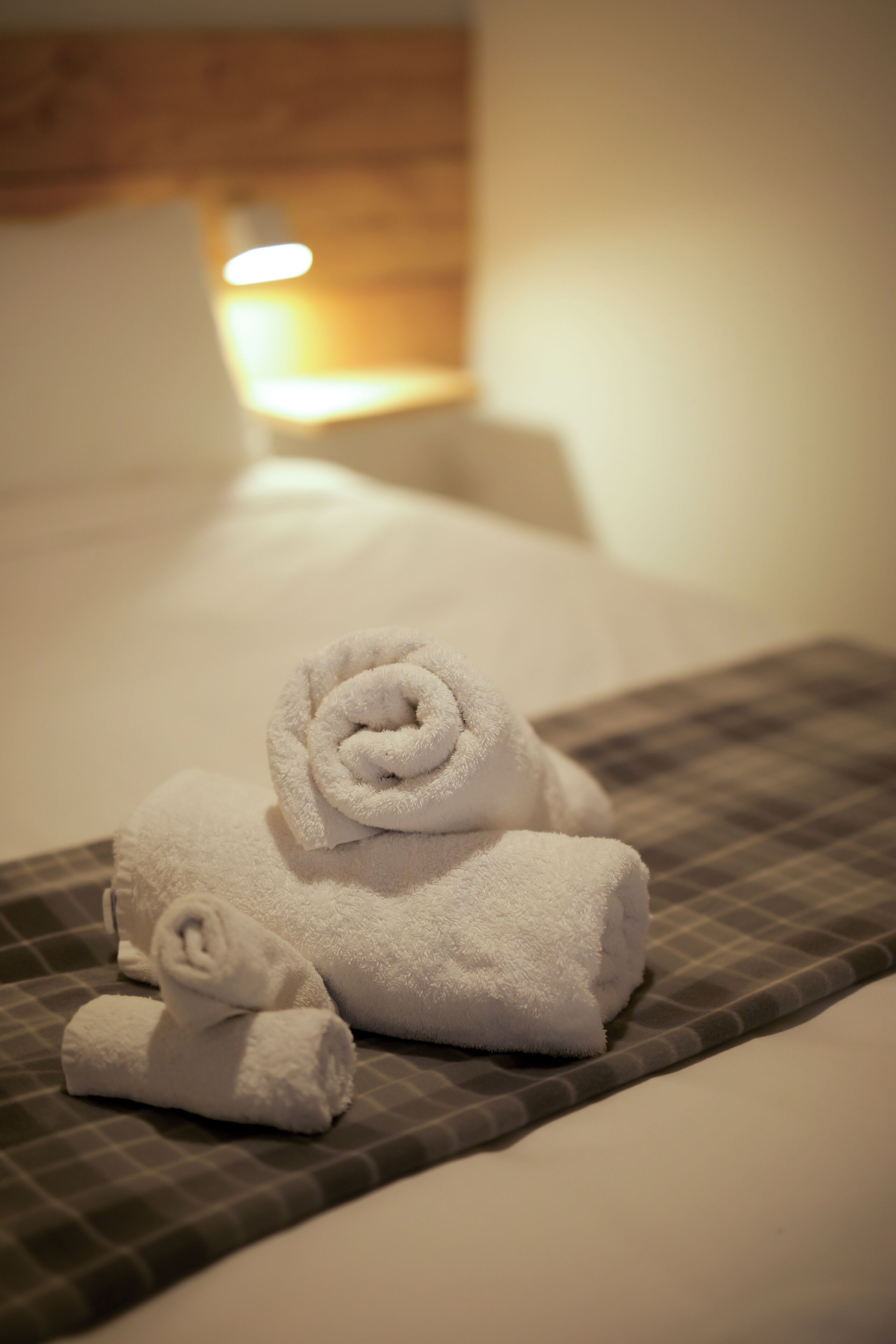 A neatly made bed with two rolled towels and a pillow, a checkered bedspread, and a bedside lamp.