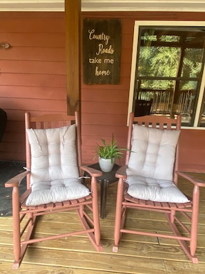 Two rocking chairs with cushioned seats and backs, positioned on a wooden deck with a potted plant between them. A sign on the wall reads "Country Roads take me home."