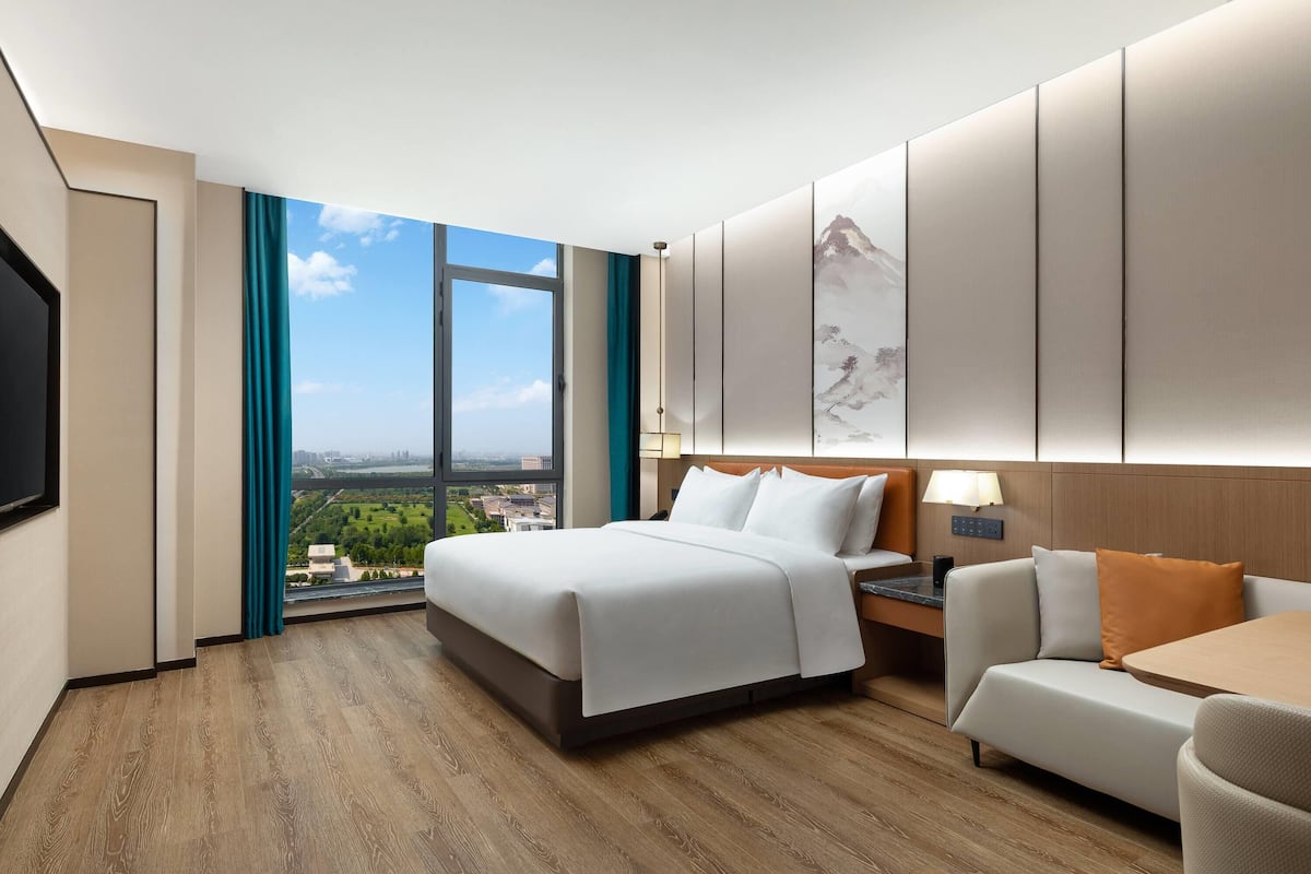 Una habitación de hotel con una cama grande, un sofá, una mesa de comedor y vistas a la ciudad a través de un ventanal amplio.