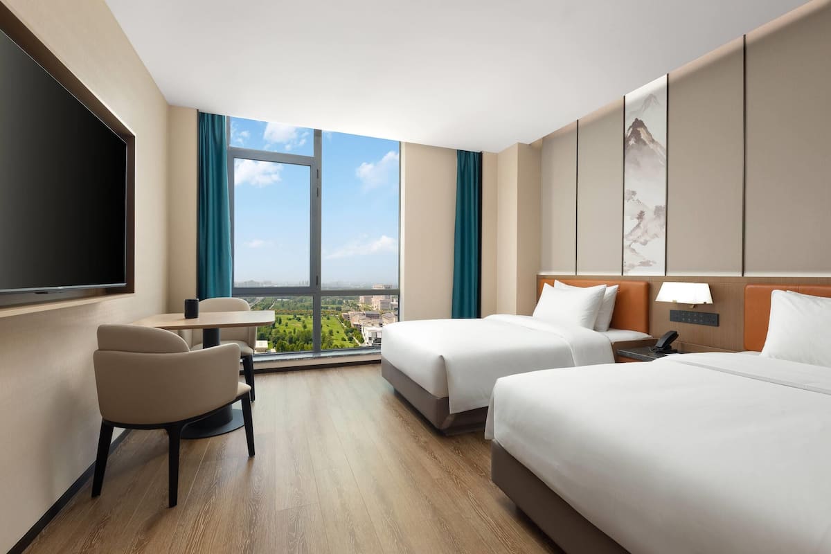 Habitación de hotel con dos camas, un escritorio, una silla, un televisor y un ventanal con vistas a la ciudad.
