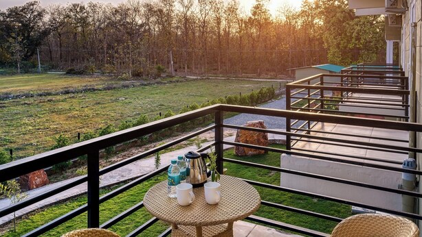 Uma varanda com móveis de vime, uma mesa com duas chávenas e garrafas, e vista para um campo gramado e árvores.