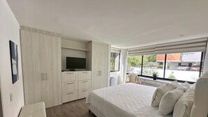 Een moderne slaapkamer met een groot bed, een televisie op een inbouwmeubel, en uitzicht op de straat door een raam.