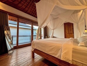 A bedroom with a wooden bed, white bedding, and a canopy. There is a sliding glass door leading to a balcony.