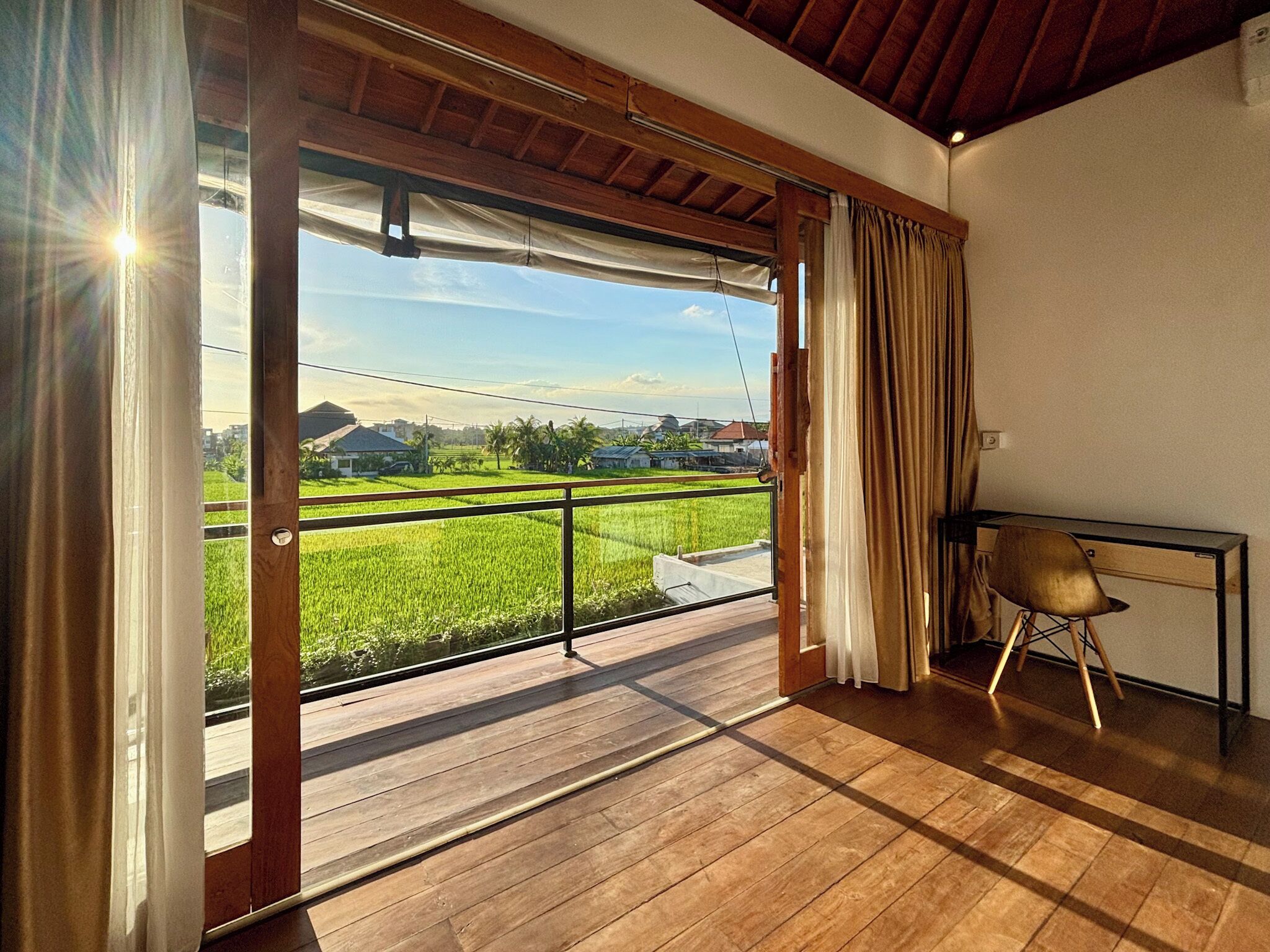A room with a wooden floor, a table with a chair, and a view of a green field through a sliding door.