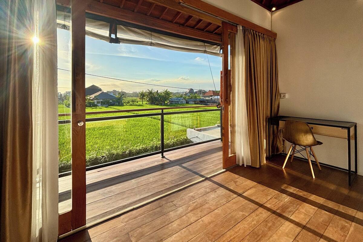 A room with a wooden floor, a table with a chair, and a view of a green field through a sliding door.
