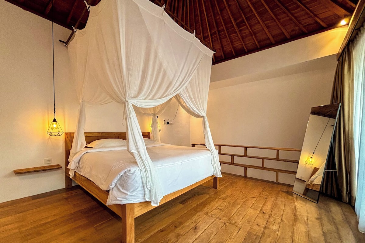 A room with a wooden bed, white canopy, wooden floor, and a small shelf.