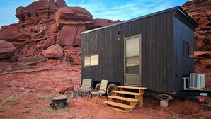 A small, dark wooden cabin with a large sliding door, a small porch with a chair and a table, and an air conditioning unit. - Cozy Cabin @ Private Moab Ranch (Moab)